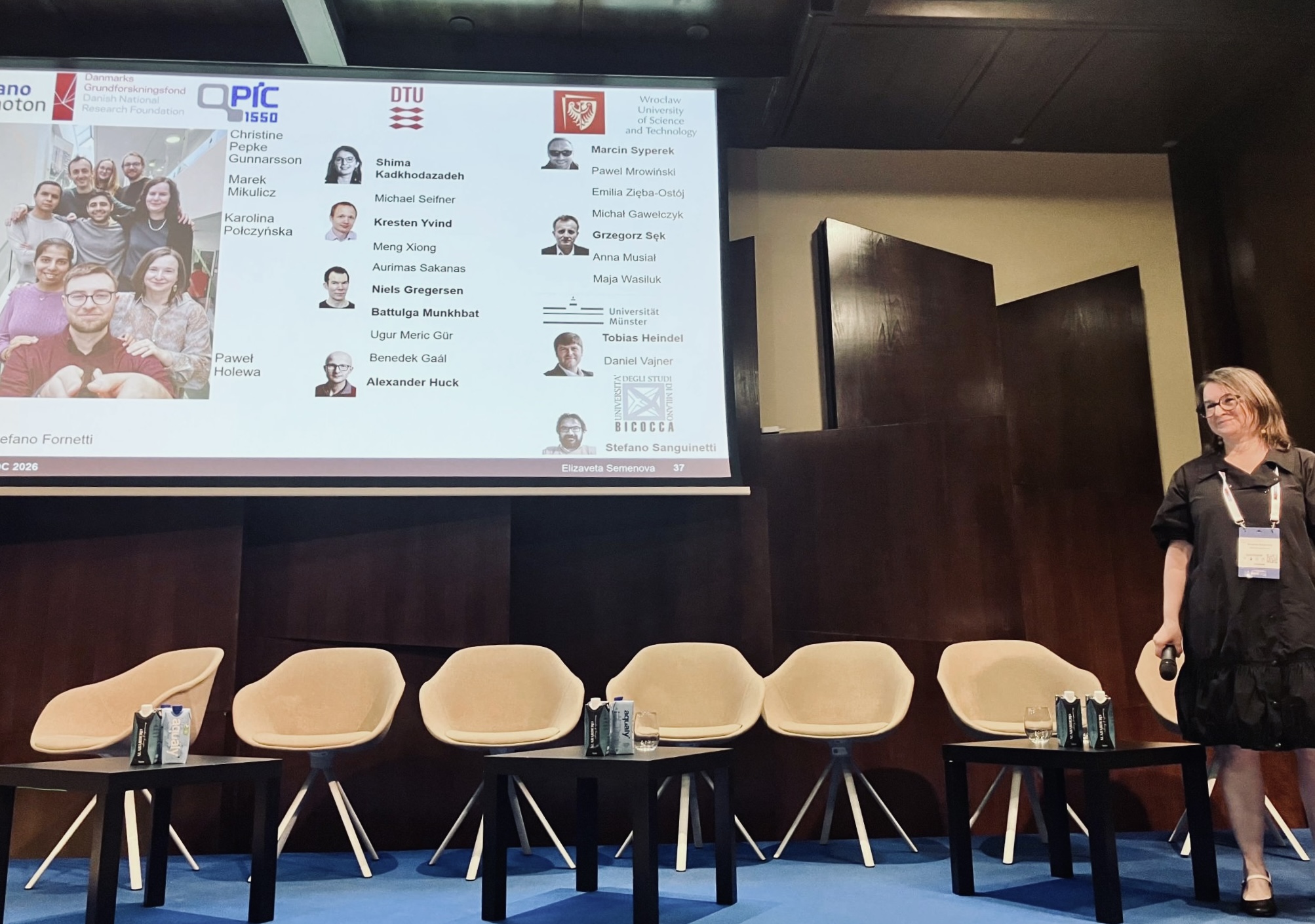 Prof. Elizaveta Semenova stands on stage holding a microphone next to a slide acknowledging project collaborators and team members from institutions like DTU.