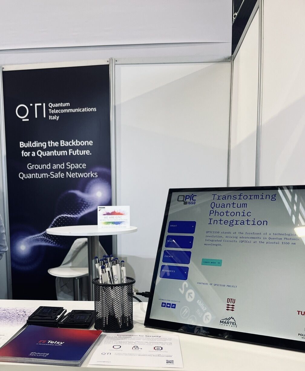 Exhibition booth for Quantum Telecommunications Italy featuring promotional flyers, pens, and a computer monitor displaying "Transforming Quantum Photonic Integration"
