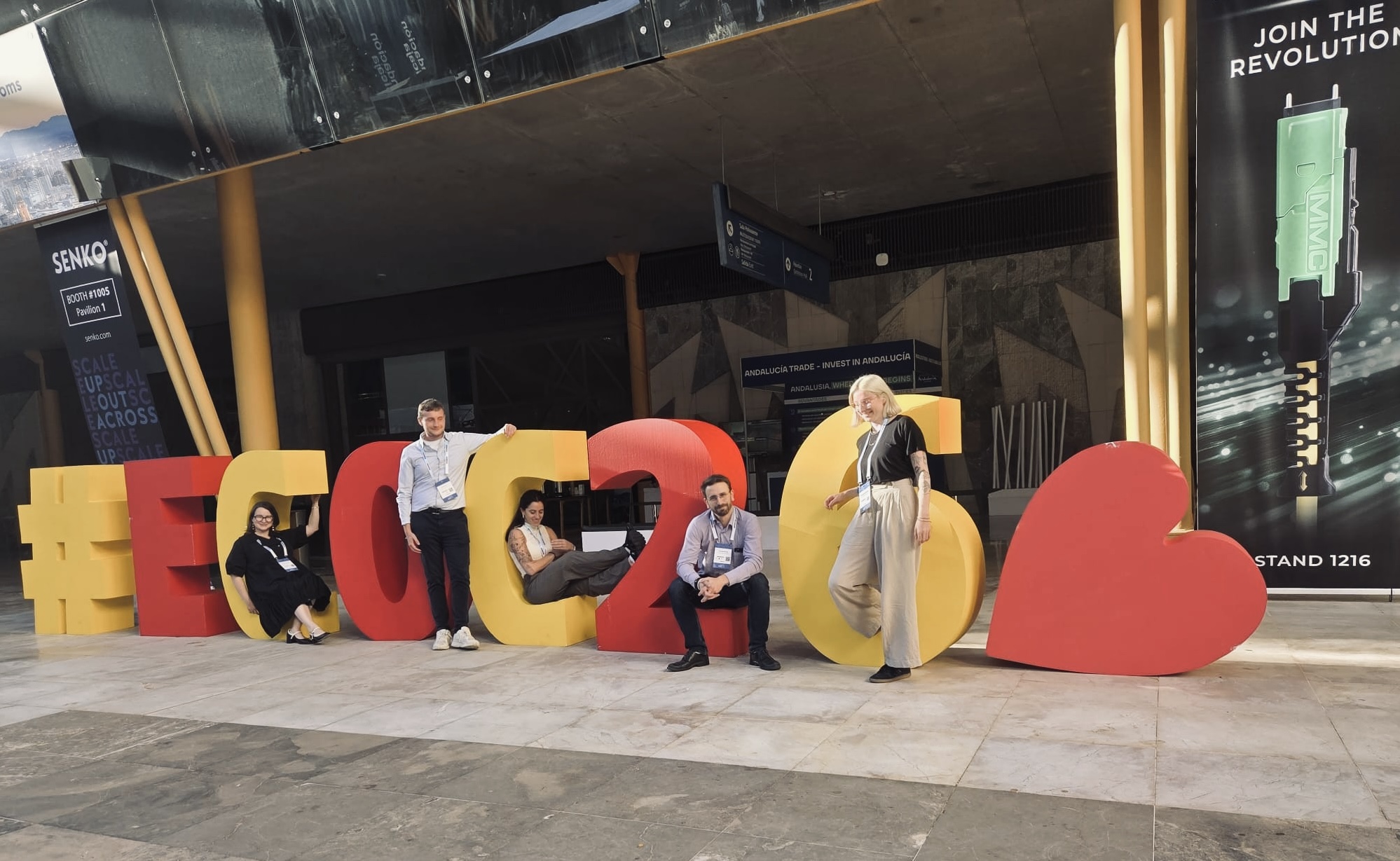 Five people pose around large, colorful 3D letters spelling out "#ECOC26" followed by a red heart symbol inside a modern exhibition hall.